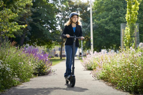 Woman in jeans, blue blazer, and helmet riding electric scooter on pathway with flowers and trees in background